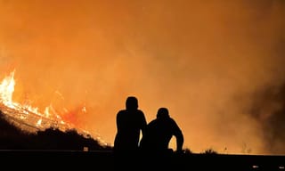 Two people silhouetted against an orange sky watching a wildfire burn across a hillside