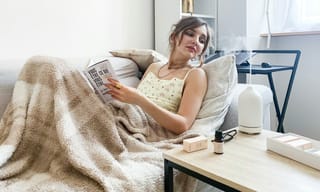 A person relaxing on a couch reading a book beside a humidifier releasing a fine mist into a cozy living room