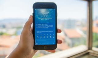 A hand holding a smartphone running a local air quality and weather app, held up to a window overlooking the city