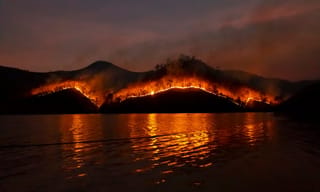 Wildfire burning across mountain ridges at night, reflected in still water below