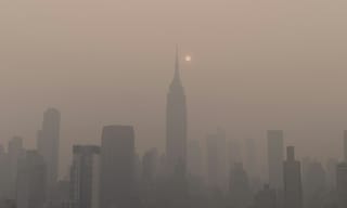 A city skyline shrouded in thick haze during wildfire season, with the sun dimmed to a pale disc behind the smoke