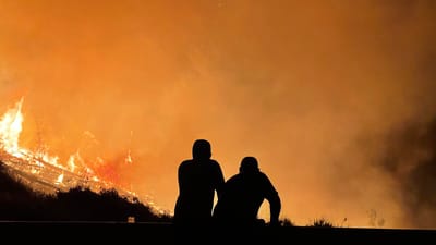 Two people silhouetted against an orange sky watching a wildfire burn across a hillside