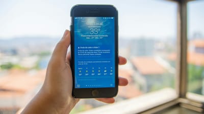 A hand holding a smartphone running a local air quality and weather app, held up to a window overlooking the city