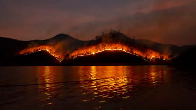Wildfire burning across mountain ridges at night, reflected in still water below