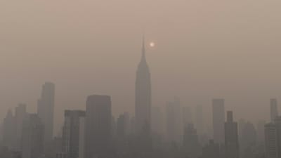 A city skyline shrouded in thick haze during wildfire season, with the sun dimmed to a pale disc behind the smoke