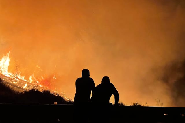 Two people silhouetted against an orange sky watching a wildfire burn across a hillside