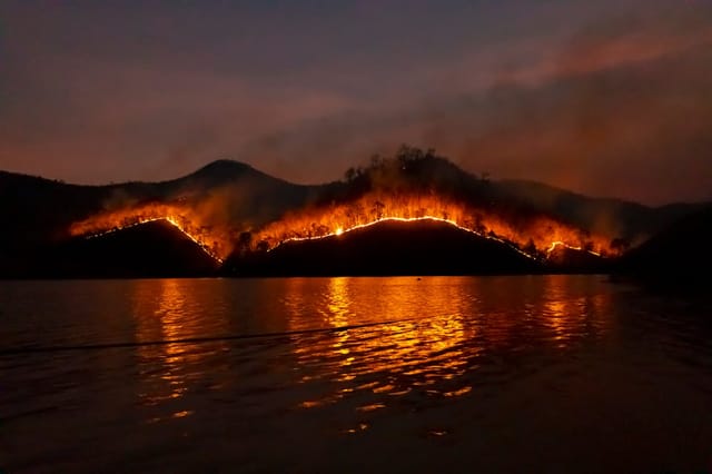 Wildfire burning across mountain ridges at night, reflected in still water below