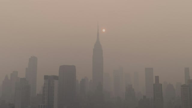 A city skyline shrouded in thick haze during wildfire season, with the sun dimmed to a pale disc behind the smoke